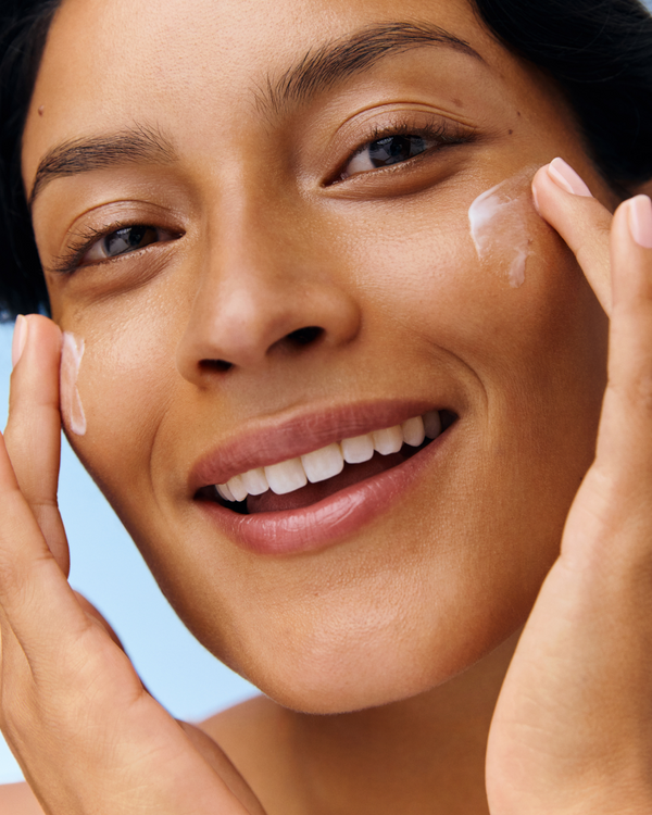 Close-up of a smiling woman applying white cream to both cheeks with her fingertips.