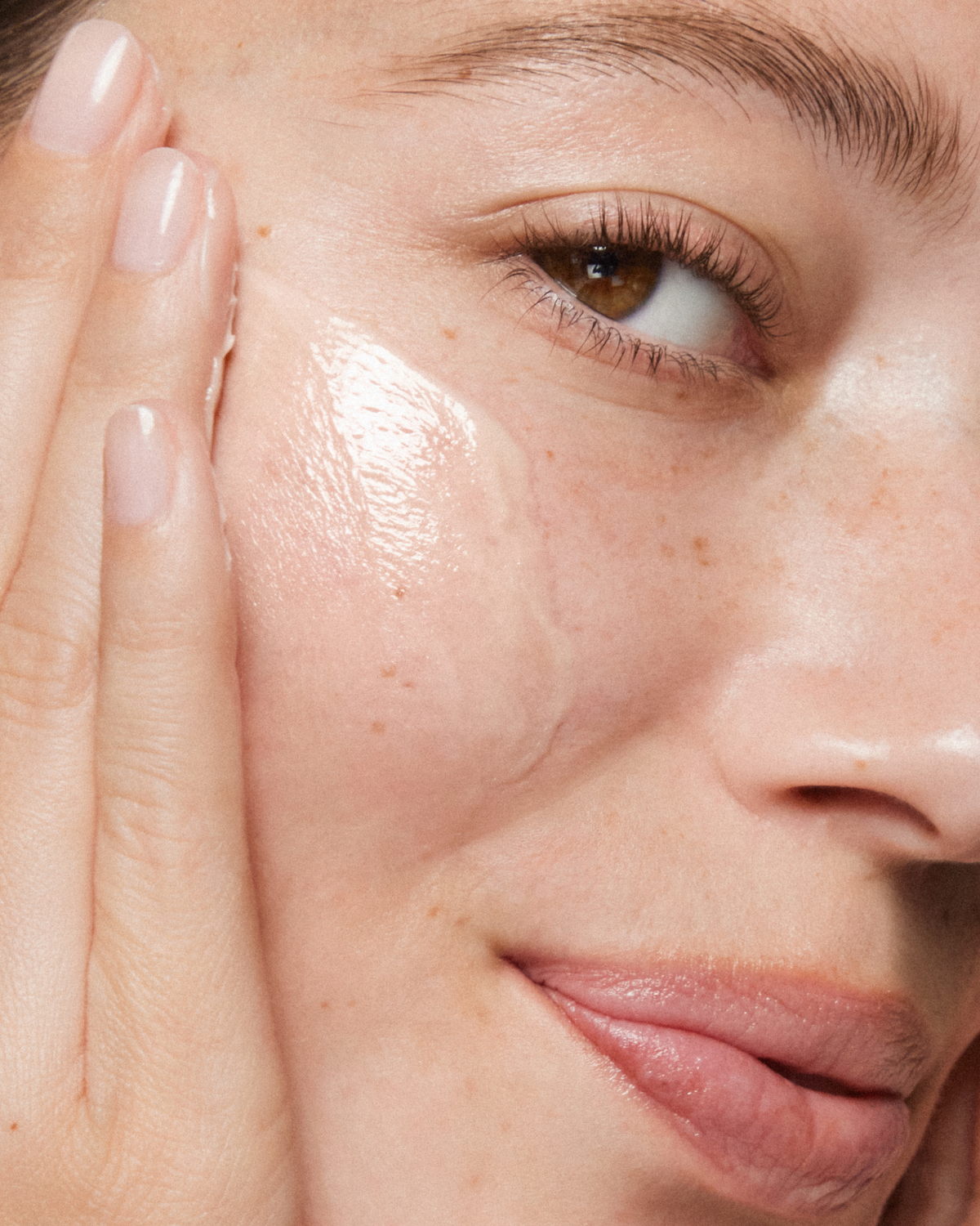 A close-up of a person applying a serum to their cheek looking into camera